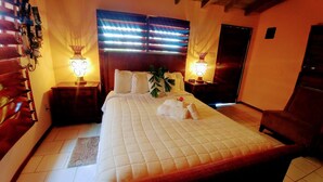 A hotel room with a large bed, two bedside tables with lamps, a potted plant, and a chair. - Yocamatsu (Caye Caulker)