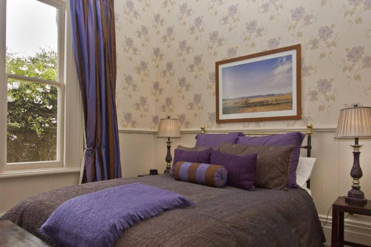 Une chambre à coucher avec un grand lit, un papier peint à motifs floraux, un tableau représentant un paysage encadré et une fenêtre avec des rideaux.