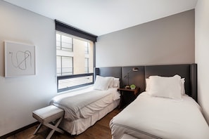 A modern hotel room with two beds, a small bench, a nightstand with a lamp, and a window with blinds. - ATTIK SUPERIOR+BALCONY 402 (Bogotá)