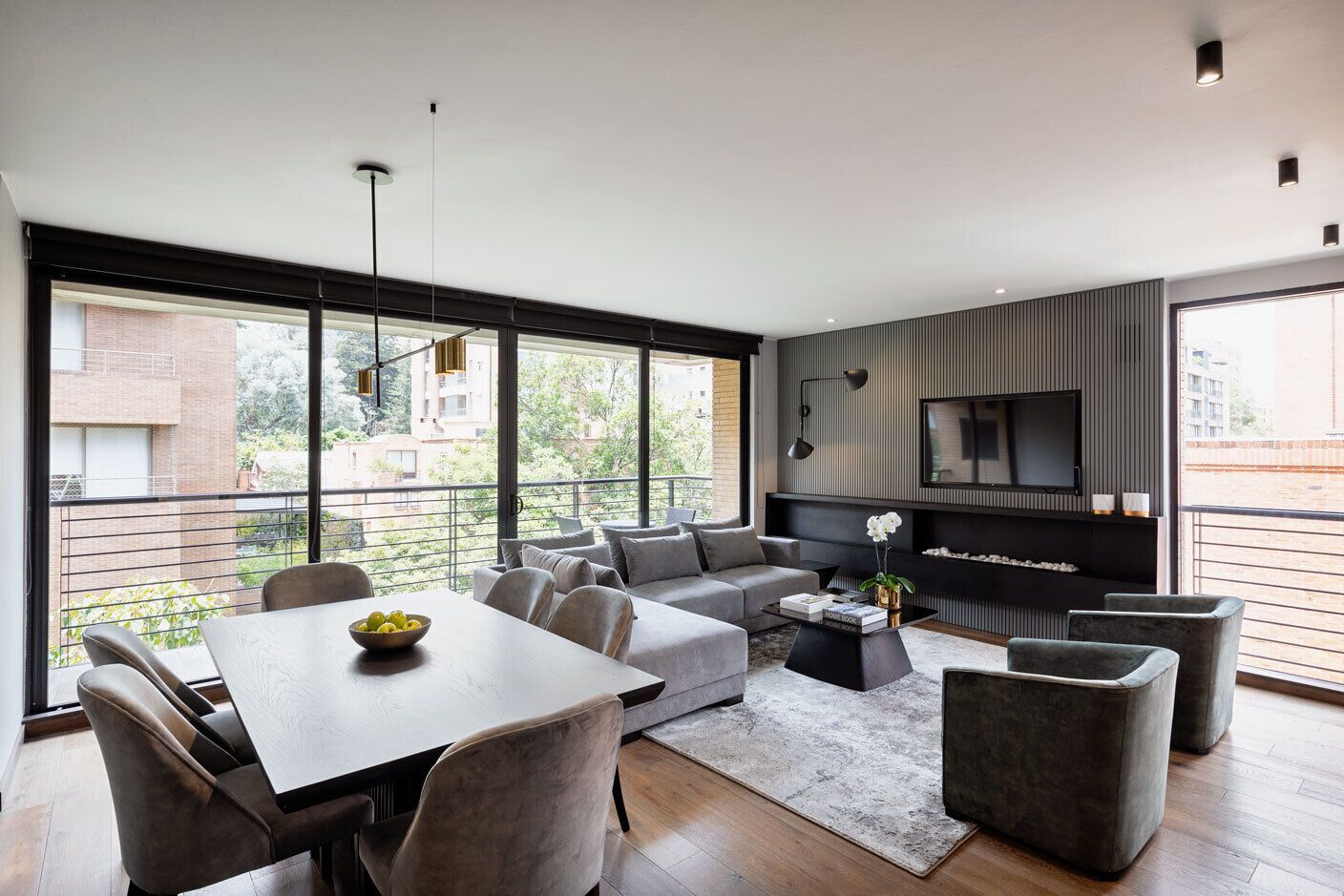En moderne stue med sofa, sofabord og spisebord. Et fjernsyn er monteret på væggen, og der er en balkon med gelænder.