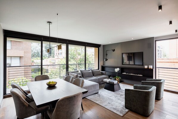 A modern living room with a sofa, coffee table, and dining table. There is a TV mounted on the wall and a balcony with a railing. - ATTIK SUPERIOR+BALCONY 402 (Bogotá)
