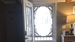 Un couloir avec un tapis à motifs, une porte fermée ornée d’un panneau de verre décoratif et une lampe posée sur une commode.