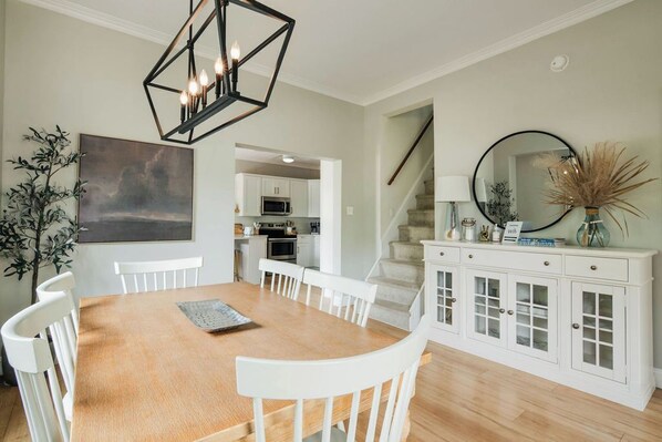 Een eetgedeelte met een houten tafel en witte stoelen, een wit kastje met glazen deuren, een ronde spiegel en een geometrische kroonluchter.
