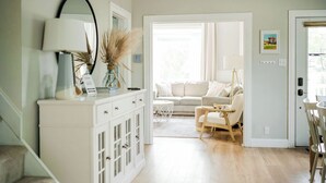 A bright, well-lit living room with a white sofa, a small table, and a chair. There is a staircase on the left and a door on the right. - Remodeled Home Near Beach and Downtown (Manistee)