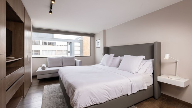 A modern bedroom with a large bed, a sofa, a TV, and a window with a city view. - NEW DESIGN DUPLEX - BOGO64 303 (Bogotá)