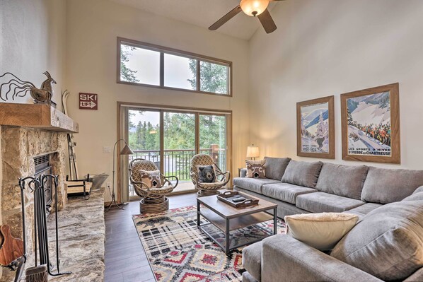 A spacious living room with a fireplace, a large window, a ceiling fan, and two framed pictures on the wall. - Walk to Lift: Townhome w/ Shuttle in Breckenridge! (Breckenridge)