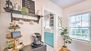 Eine Küche mit einer Kaffeestation, Regalen voller Küchenutensilien, einem blauen Schrank und einem Fenster mit Jalousien.