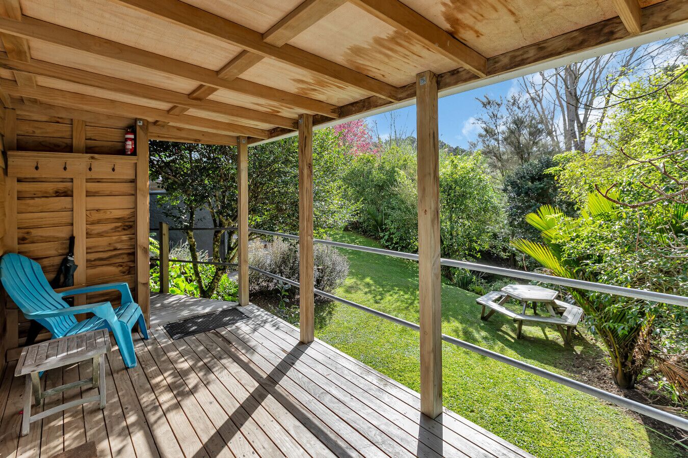 Un terrazzo in legno con zona coperta, una sedia blu, un tavolo e vista su un giardino con tavolo da picnic.