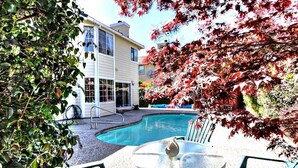 A swimming pool with lounge chairs and a table, surrounded by a building with large windows and a garden with autumn foliage. - Grand Executive Luxury Home (Union City)