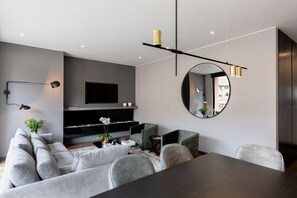A modern living room with a grey sofa, a coffee table, and a TV mounted on the wall. There are two chairs, a round mirror, and a dining table. - ATTIK TOP OF THE TOP - 602 (Bogotá)