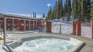 Ett område med ett bubbelbad och en pool, omgivet av en röd byggnad med ett snötäckt staket och träd i bakgrunden.