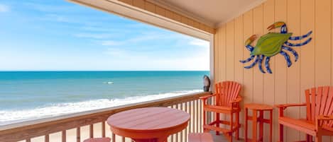 Una terraza de madera con una mesa redonda y seis sillas, con vista a una playa y una vista despejada del océano.