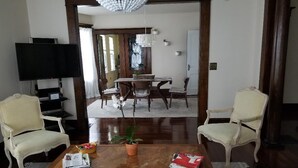 A living room with a wooden coffee table, two armchairs, a TV, and a dining area with a chandelier.