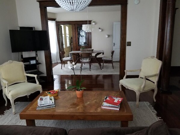 A living room with a wooden coffee table, two armchairs, a TV, and a dining area with a chandelier.