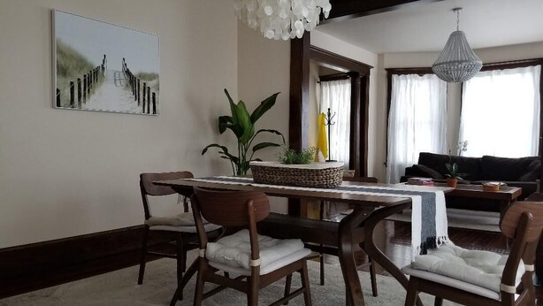 A dining area with a long table, four chairs, a chandelier, and a painting on the wall.