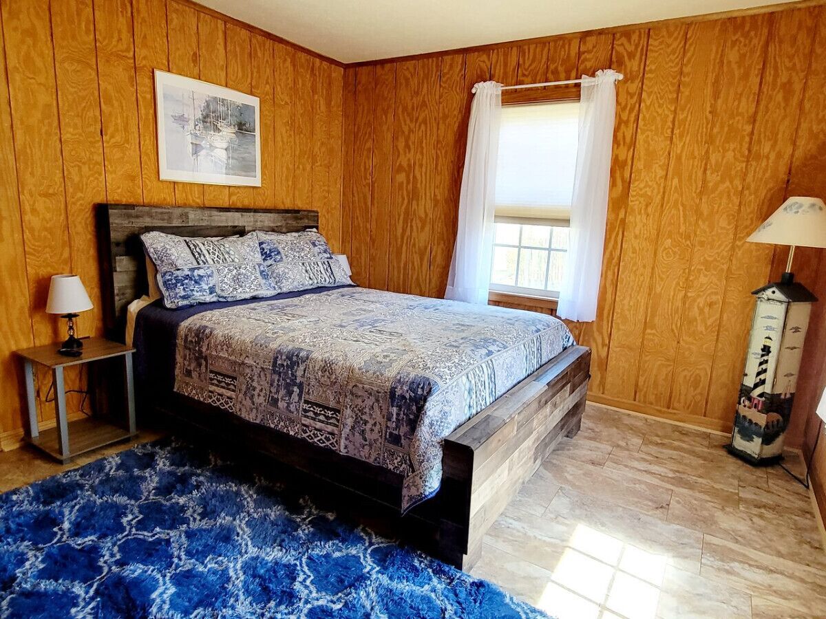 A bedroom with a wooden paneled wall, a bed with patterned bedding, a nightstand with a lamp, and a window with curtains.