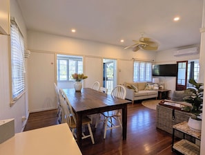 A spacious room with a wooden dining table, white chairs, a sofa, a television, and a ceiling fan.