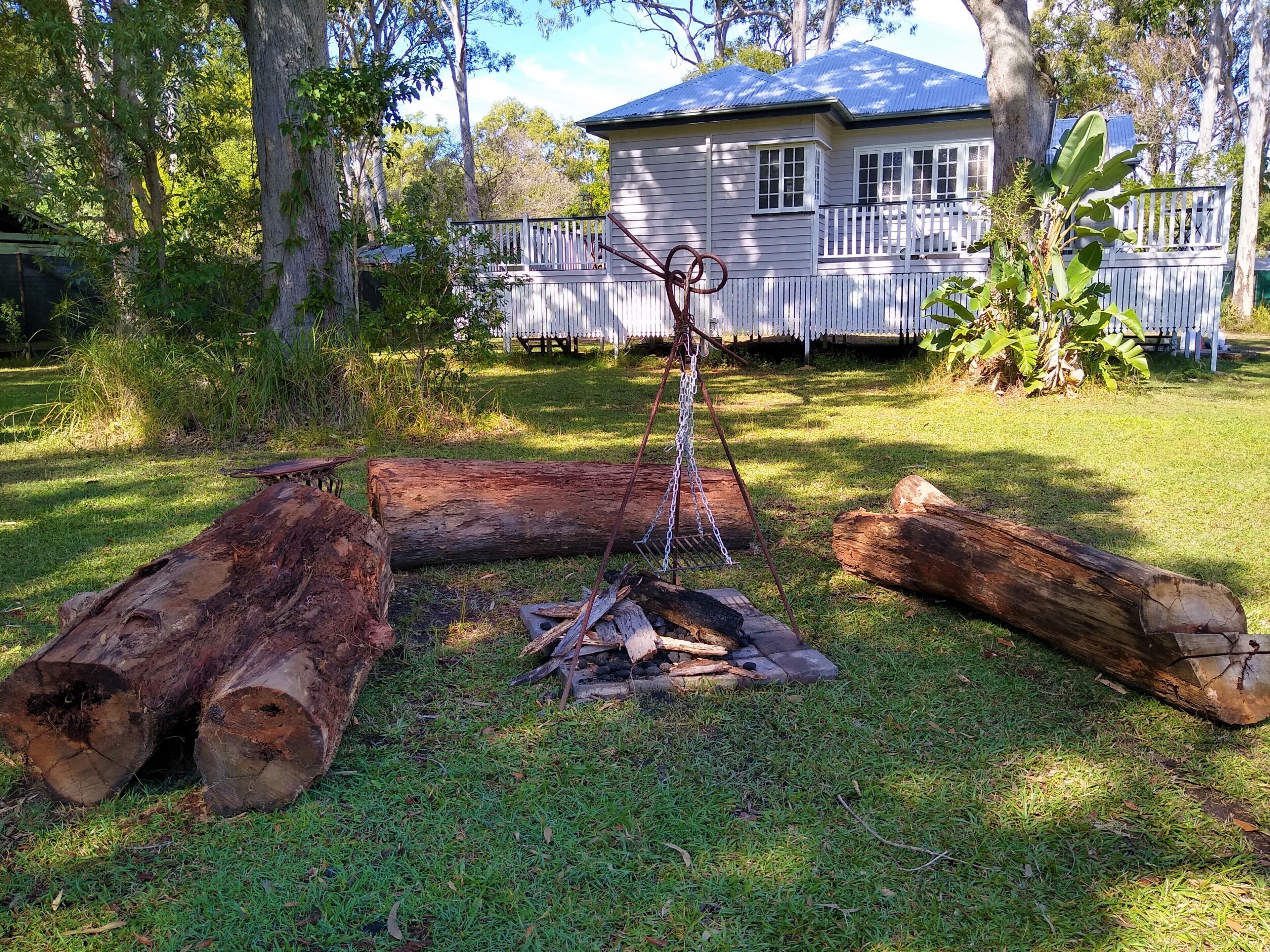 A backyard with a fire pit surrounded by logs, a white fence, and a house with a blue roof in the background.