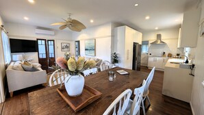Une cuisine moderne avec des meubles blancs, un coin repas doté d’une table en bois et de chaises blanches, et un ventilateur de plafond.