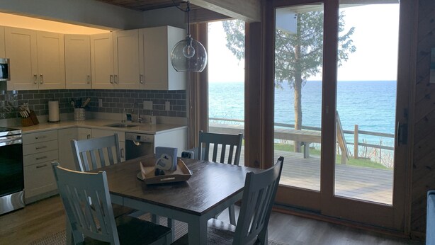 Una cocina moderna con armarios blancos, una mesa de comedor de madera y sillas. Un ventanal ofrece vistas al mar.
