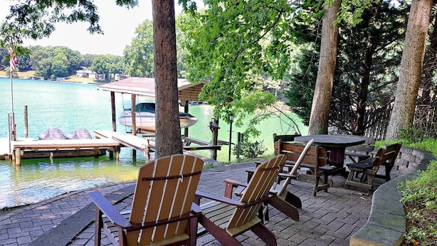 Une terrasse au bord de l’eau, avec des fauteuils Adirondack et une table, d’où l’on peut admirer un lac avec des bateaux et un quai.
