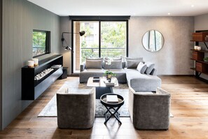 Uma sala de estar moderna com um sofá seccional, duas poltronas, uma mesa de centro e uma lareira. Há uma janela grande com vista para o verde lá fora.