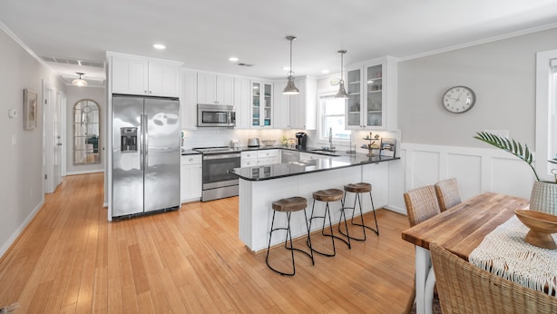 A modern kitchen with white cabinetry, stainless steel appliances, a central island with bar stools, and a dining area with a wooden table and wicker chairs. - OBX Beach Box!  204 E Albatross St Milepost 10 (Nags Head)