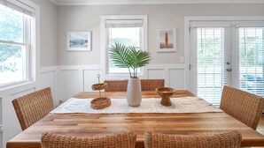 A dining area with a wooden table, wicker chairs, a central vase with a plant, and framed pictures on the walls. - OBX Beach Box!  204 E Albatross St Milepost 10 (Nags Head)