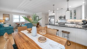 A modern kitchen with white cabinets, stainless steel appliances, and a wooden dining table with wicker chairs. - OBX Beach Box!  204 E Albatross St Milepost 10 (Nags Head)