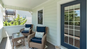 A covered porch with wicker chairs and a small table, a large window, and a view of greenery. - OBX Beach Box!  204 E Albatross St Milepost 10 (Nags Head)