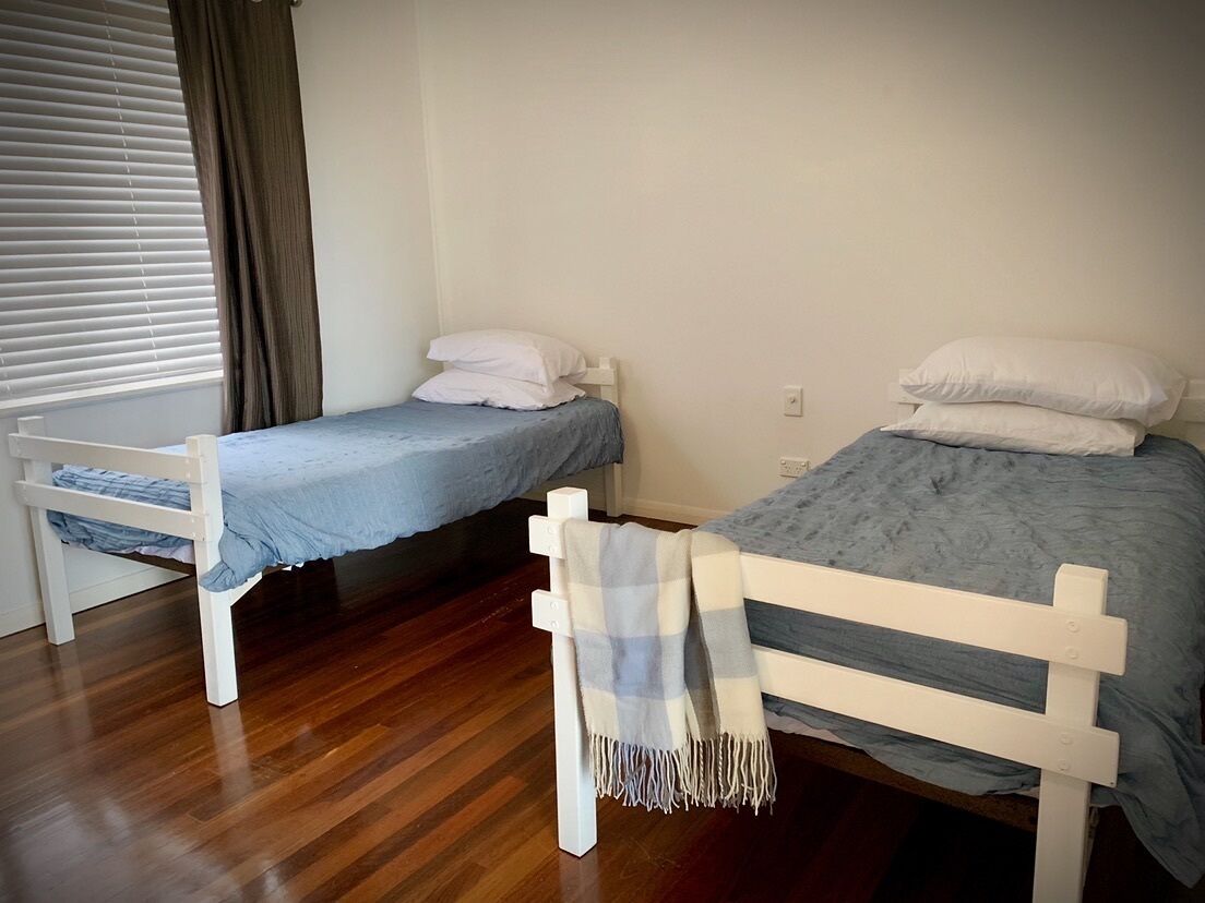 Two single beds with white frames and blue bedding, a wooden floor, and a window with blinds and a curtain.