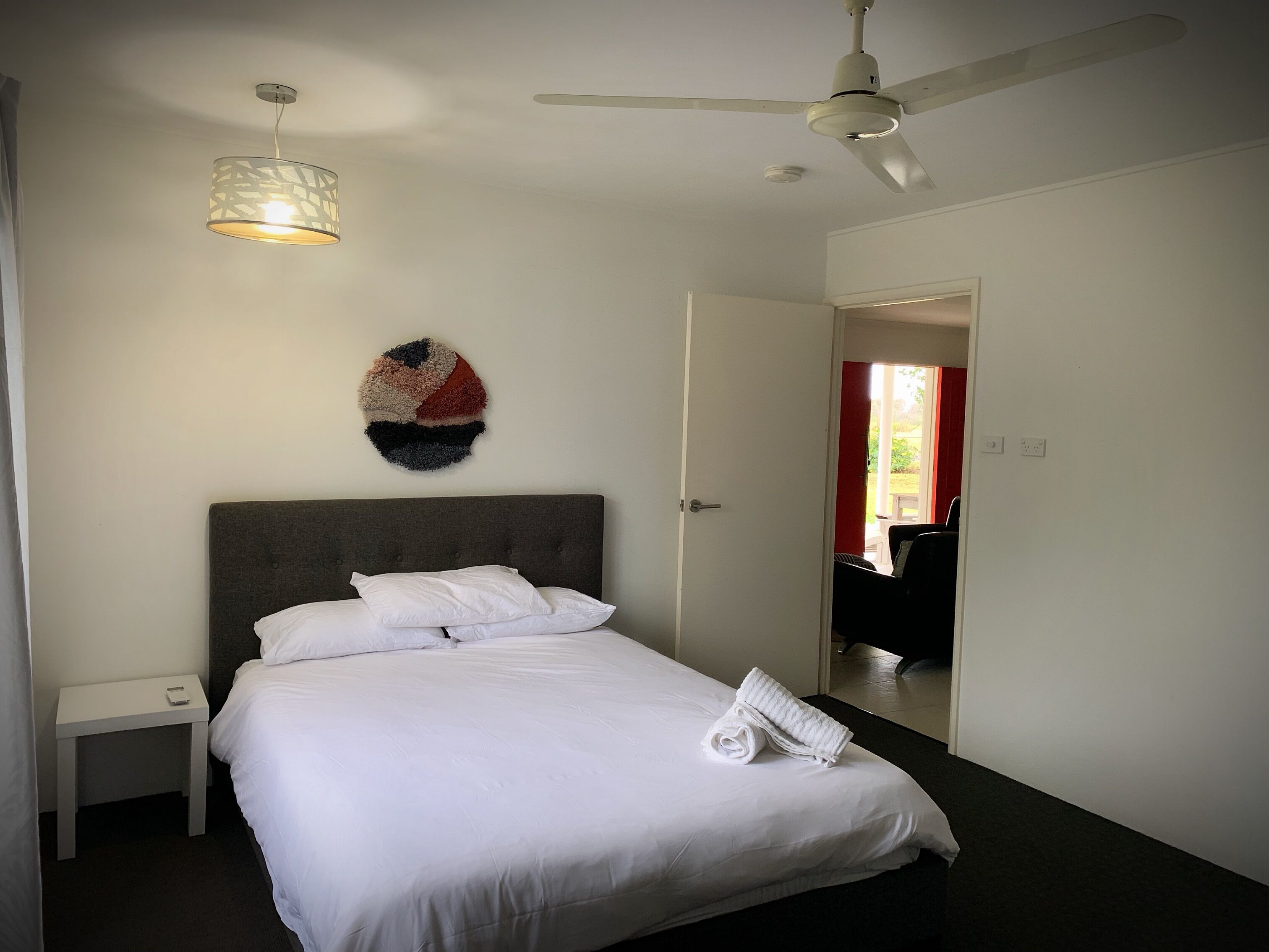 A neatly made bed with white linens, a bedside table, a wall-mounted decorative piece, and a ceiling fan.
