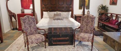 Una cama con dosel, un tapete con estampado floral, dos sillones, un cofre de madera y un sofá rojo.