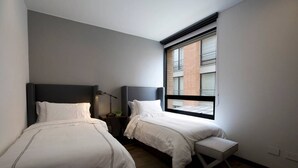 Habitació d'hotel moderna amb dos llits, una banca, una tauleta de nit i una finestra amb vista a un edifici de maó.