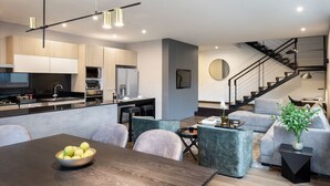 Una cuina moderna amb una illa central, una zona de menjador amb taula i cadires de fusta, i una sala d'estar amb sofà i taula de centre.