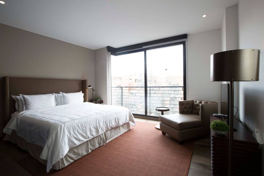 A modern hotel room with a large bed, a small sofa, a desk, and a large window offering a city view.