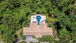 An aerial view of a resort with a central pool, surrounded by lush greenery and palm trees.
