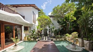 A pool with a wooden deck, surrounded by potted plants and a white building with large windows.