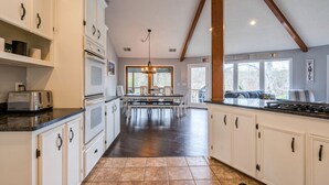 A spacious kitchen with white cabinetry, a dining area with a large table, and a view of the outdoors through the windows.