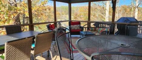 A covered outdoor seating area with metal tables and wicker chairs, overlooking a forested area.