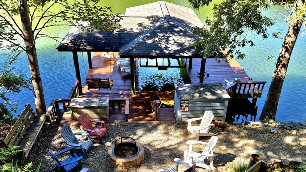 A deck with Adirondack chairs, a fire pit, and a covered seating area overlooking a lake.