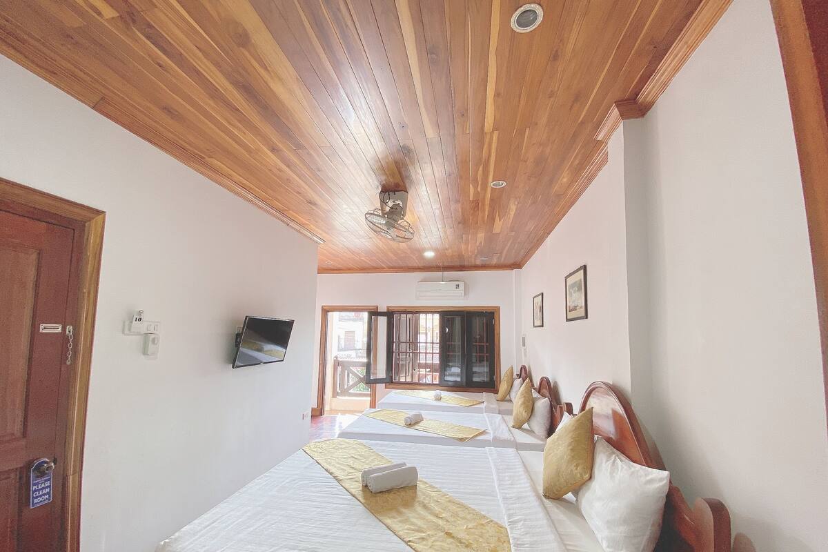 A hotel room with wooden ceiling beams, two beds with white linens and decorative pillows, a flat-screen TV mounted on the wall, and a sliding glass door leading to an outdoor area.