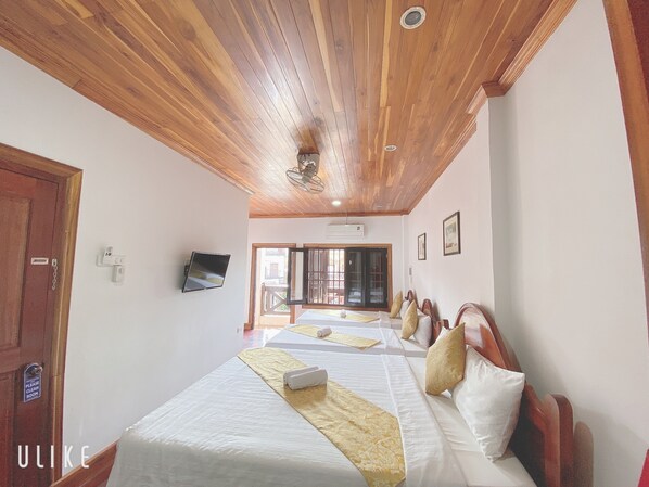 A hotel room with wooden ceiling beams, two beds with white linens and decorative pillows, a flat-screen TV mounted on the wall, and a sliding glass door leading to an outdoor area.