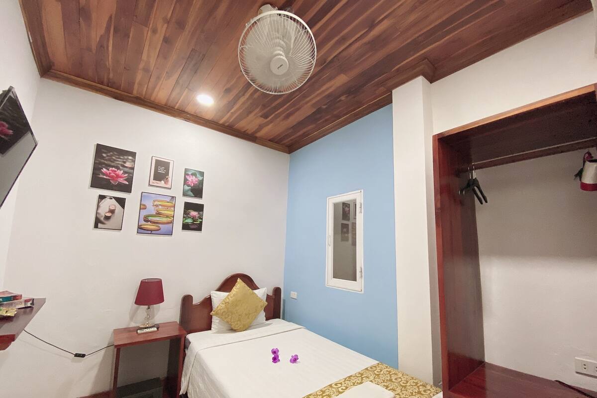 A bedroom with a wooden ceiling, a bed with a white bedspread, a bedside table with a lamp, and a wall-mounted fan.
