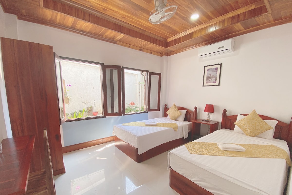 A hotel room with two beds, wooden ceiling, large windows, and a bedside table with a lamp.