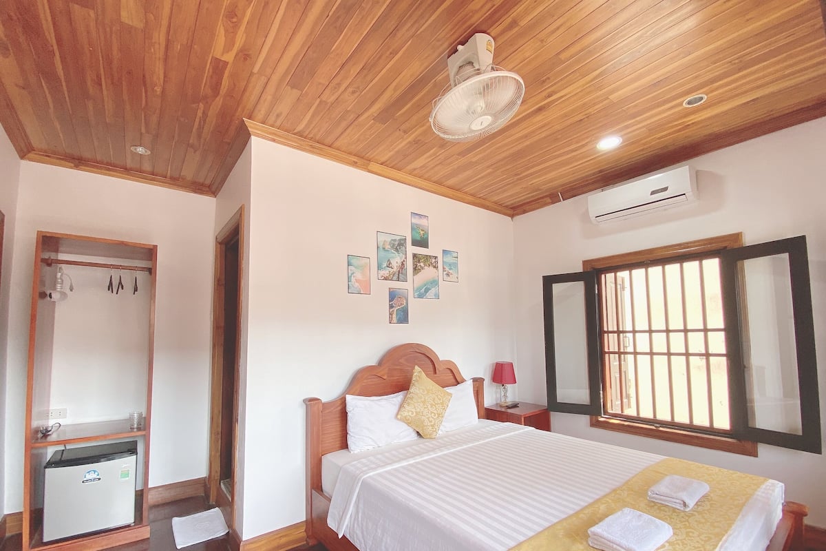 A bedroom with a wooden ceiling, a bed with white bedding, a small bedside table with a lamp, and a window with bars.
