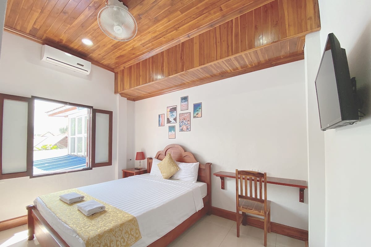 A hotel room with a wooden ceiling, a bed with a white and gold bedspread, a wooden nightstand with a lamp, a wooden chair, and a TV mounted on the wall.