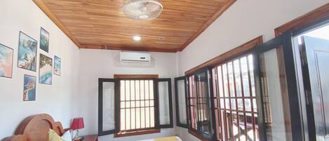 Habitación de hotel con techo de madera, cama con ropa blanca, lámpara roja y ventana con barrotes.
