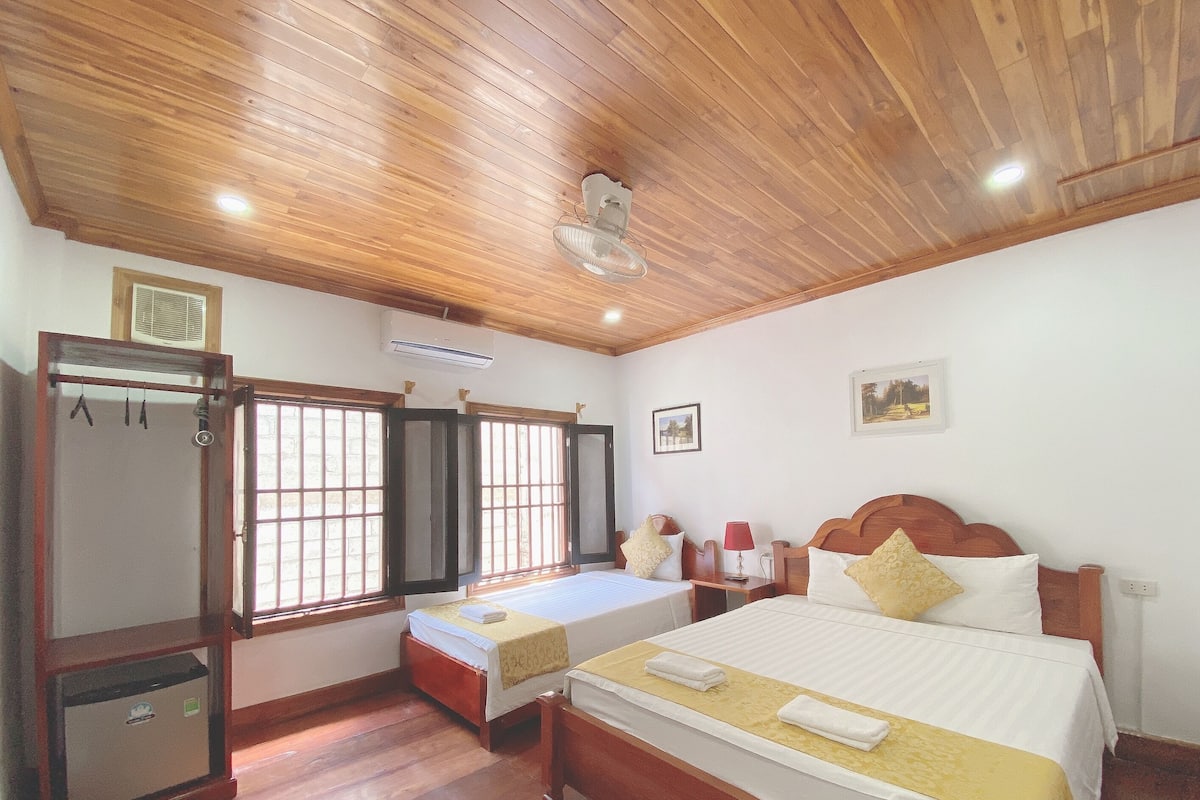 A hotel room with wooden ceiling, two beds, a wardrobe, and a window with bars.