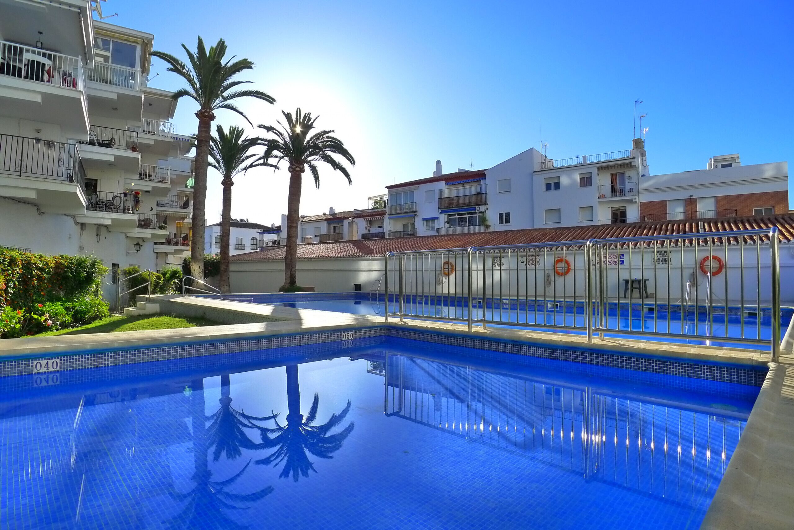 A swimming pool with clear blue water, surrounded by white buildings with balconies, palm trees, and a clear sky.